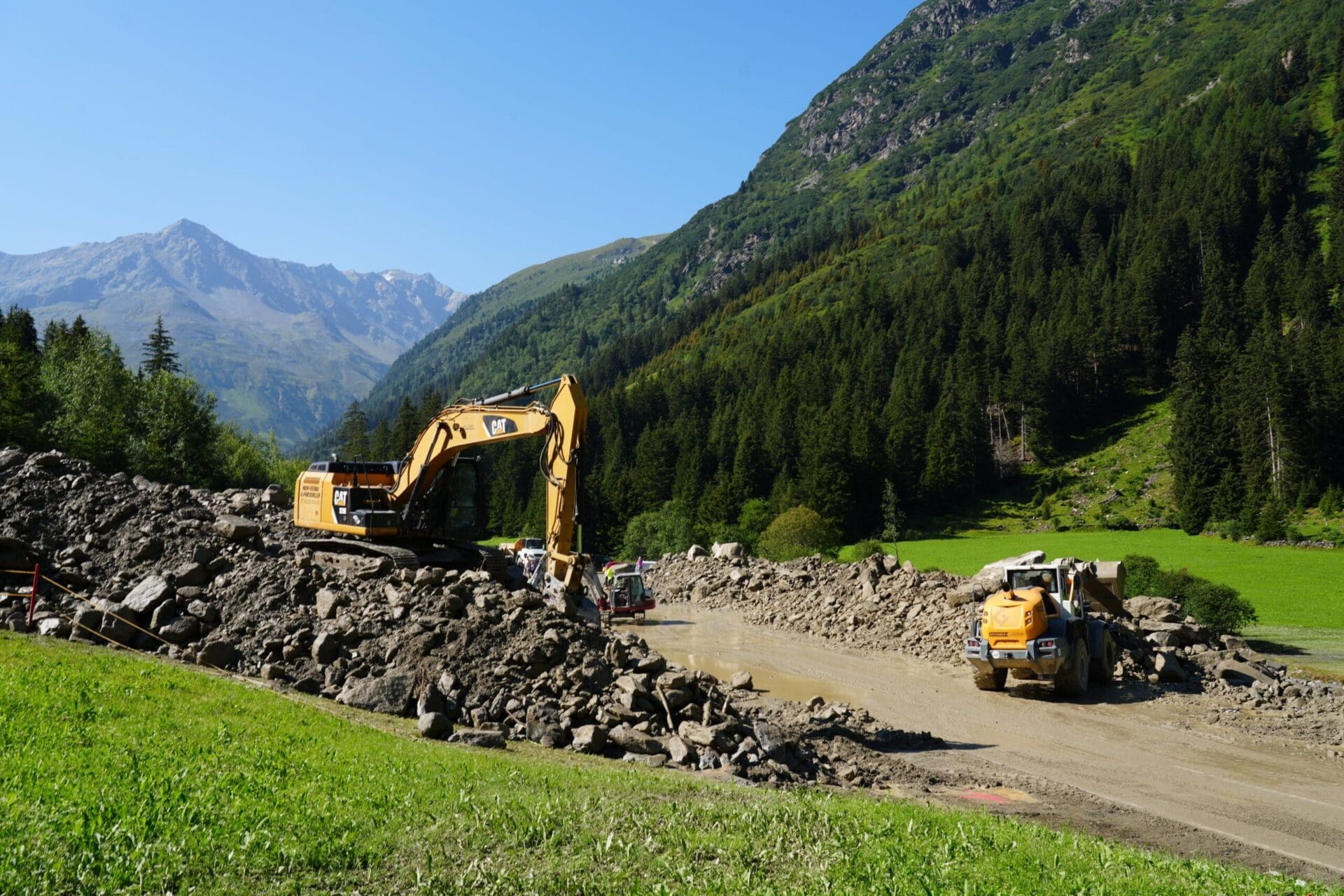 Gelber Bagger und Radlader räumen Steine und Schlamm von einer Straße in einem grünen Bergtal