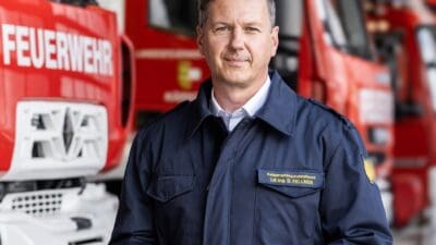 Daniel Fellner in Feuerwehruniform vor roten Einsatzfahrzeugen in einer Wache