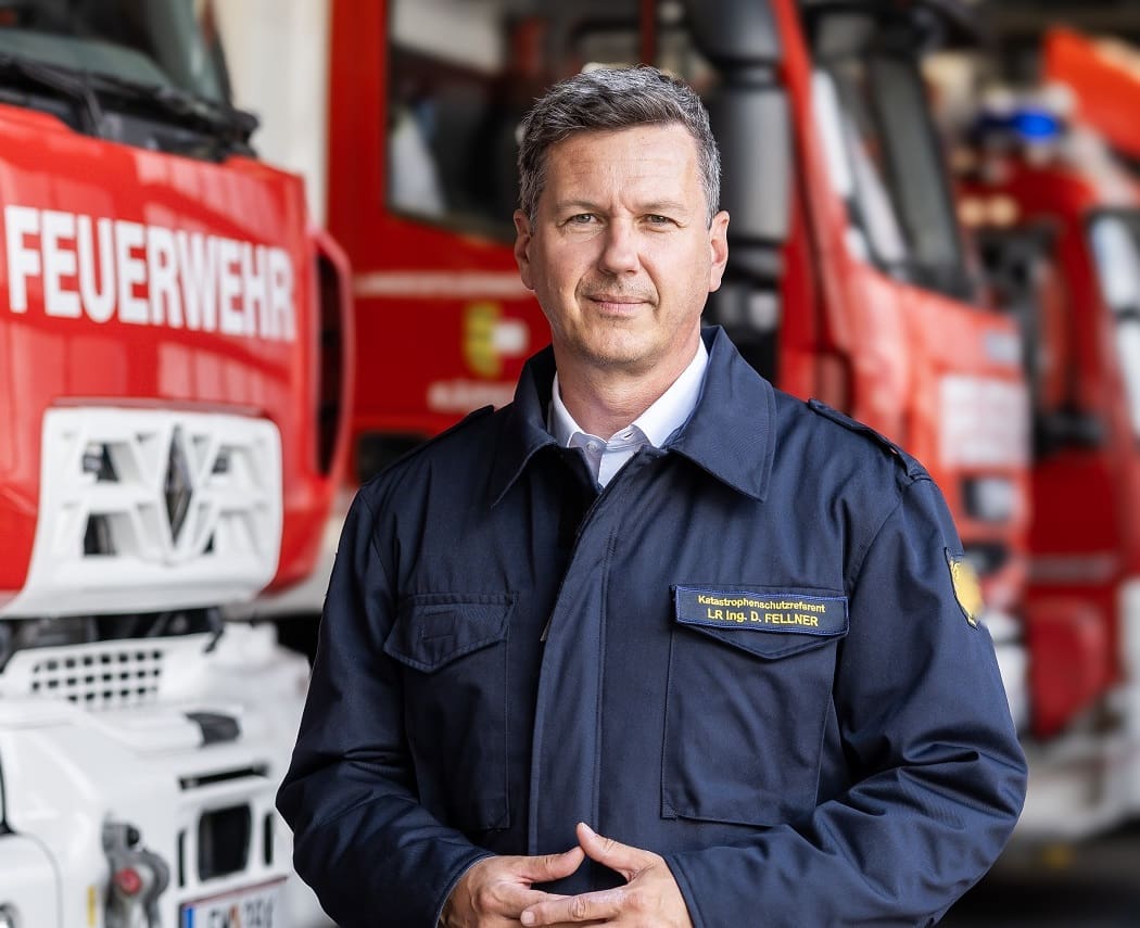 Daniel Fellner in Feuerwehruniform vor roten Einsatzfahrzeugen in einer Wache