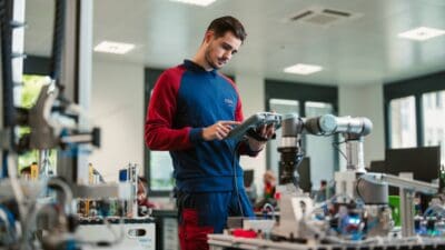 Junger Mann in ÖBB-Arbeitskleidung bedient Tablet in Werkstatt mit Industrierobotern