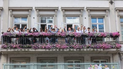 Jugendliche und Erwachsene auf Balkon mit Blumenkästen jubeln mit erhobenen Fäusten
