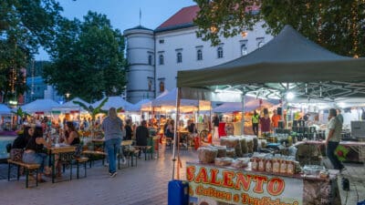 Besucher sitzen an Tischen vor einem beleuchteten Marktstand mit verpackten Lebensmitteln bei einem Fest in der Dämmerung.