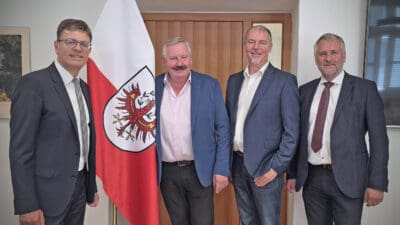 Vier Männer in Anzügen stehen nebeneinander vor einer rot-weißen Flagge mit Adler in einem Raum.