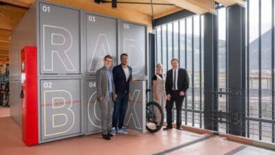 Vier Personen stehen vor grauen Radboxen mit der Aufschrift RAD BOX in einer Halle mit Ausblick auf Berge.