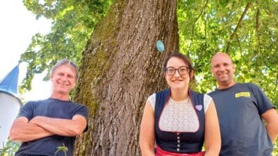 Frau in rotem Dirndl und zwei Männer vor großem Baum mit Plakette