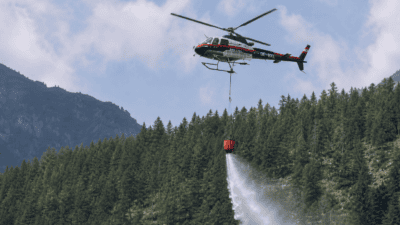 Österreichischer Polizeihubschrauber sprüht Wasser aus einem Behälter auf einen Waldbrand in den Bergen.