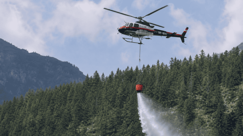 Österreichischer Polizeihubschrauber sprüht Wasser aus einem Behälter auf einen Waldbrand in den Bergen.