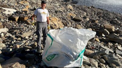 Ein Mann steht neben einem großen weißen Müllsack auf steinigem Gelände vor einem Gletscher.