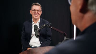 Ein lächelnder Mann in dunklem Anzug und Brille sitzt vor einem Mikrofon bei einer Podcast-Aufnahme.