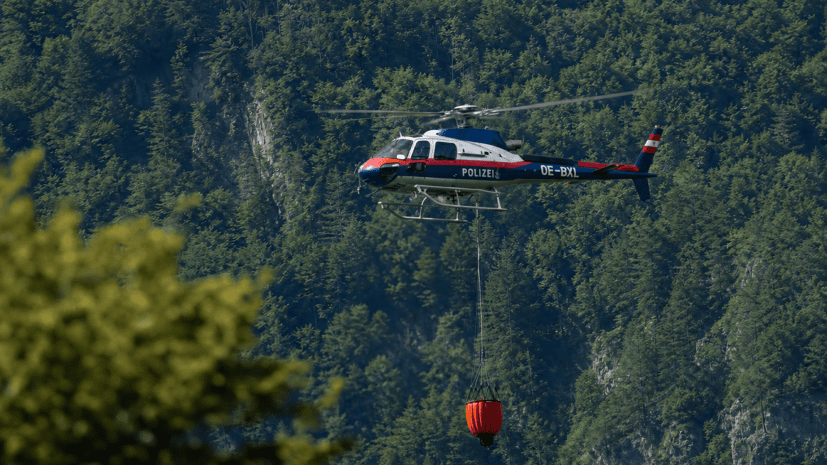 Polizeihubschrauber mit rotem Löschwasserbehälter fliegt über dicht bewaldetem Berggebiet