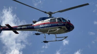 Polizeihubschrauber mit den Aufschriften POLIZEI und OE-BXL fliegt vor einem blauen Himmel mit Wolken.