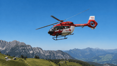 Ein roter und weißer Rettungshubschrauber der ARA Flugrettung mit Kennung OE-XFR fliegt über grünen Bergen und Tälern.