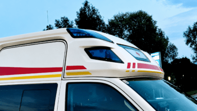 Weißer Rettungswagen mit roten und gelben Streifen sowie blauen Blinklichtern unter blauem Himmel
