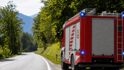 Heckansicht eines roten Feuerwehrautos mit aktiviertem Blaulicht am Rand einer Landstraße in bergiger Landschaft