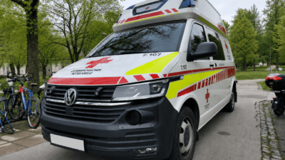 Weißer Rettungswagen des Österreichischen Roten Kreuzes mit roten und gelben Markierungen steht in einem Park.
