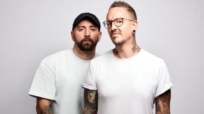 Zwei Männer mit Tätowierungen und weißen T-Shirts stehen nebeneinander vor grauem Hintergrund.