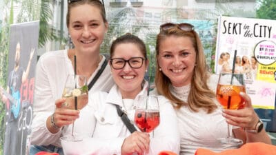 Drei Frauen lächeln in die Kamera und halten Gläser mit verschiedenen Cocktails.