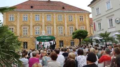 Schauspieler auf einer Buehne vor einem gelben Gebaeude mit Publikum im Vordergrund