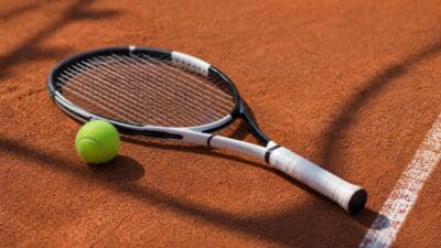 Tennisschläger mit weißem Griff neben einem gelben Tennisball auf rotem Sandplatz