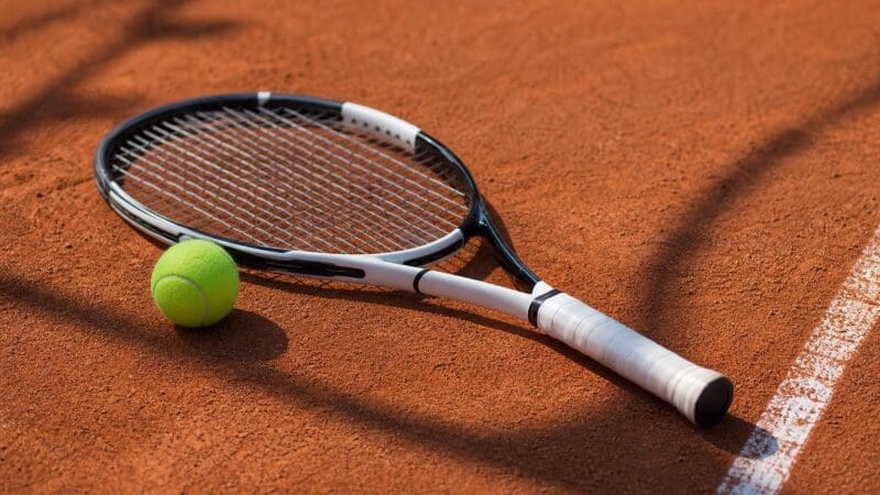 Tennisschläger mit weißem Griff neben einem gelben Tennisball auf rotem Sandplatz