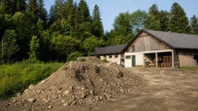 Großer Erdhaufen vor alten Holzgebäuden und Wald am Baugrundstück