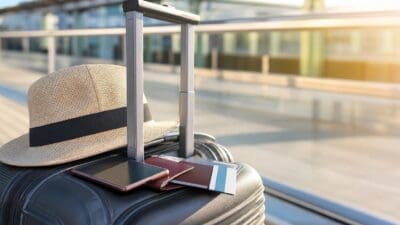 Schwarzer Rollkoffer mit Strohhut, Smartphone, Reisepass und Kreditkarte am Griff vor einem Flughafengebäude