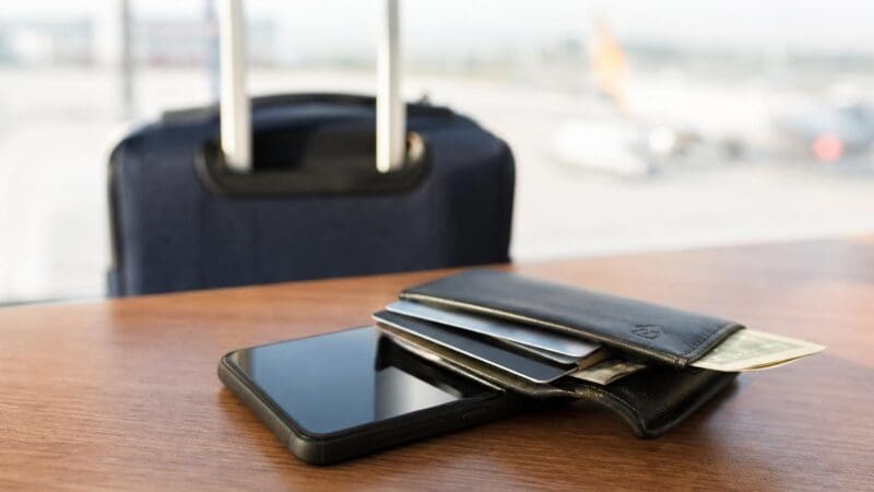 Smartphone neben schwarzer Geldbörse mit Kreditkarten und Geld auf Holztisch vor Koffer im Hintergrund