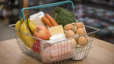Einkaufskorb mit Bananen, Äpfeln, Brokkoli, Karotten, Eiern, Käse und weiteren Lebensmitteln auf einem Holztisch