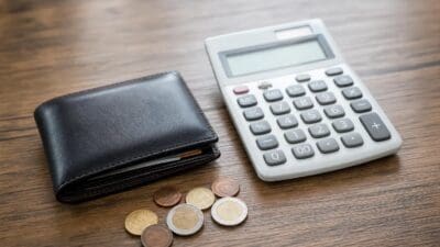 Ein schwarzes Portemonnaie liegt neben einem Taschenrechner und mehreren Euro-Münzen auf einem Holztisch.