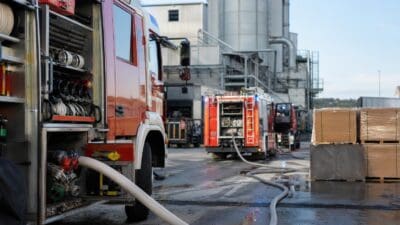Rotes Feuerwehrauto mit offenen Seitenfächern und liegenden Schläuchen auf nassem Asphalt vor Industrieanlage mit Silos