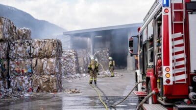 Feuerwehrleute löschen Brand von Altpapierballen vor einer Recyclinghalle mit Feuerwehrfahrzeug