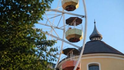 Buntes Riesenrad mit mehreren farbigen Gondeln vor gelbem Gebäude und grünen Bäumen