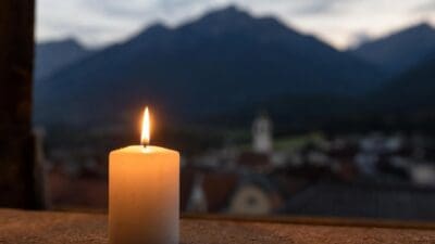 Eine brennende weiße Kerze auf einer Holzplatte vor einem verschwommenen Bergdorf mit Kirchturm im Hintergrund