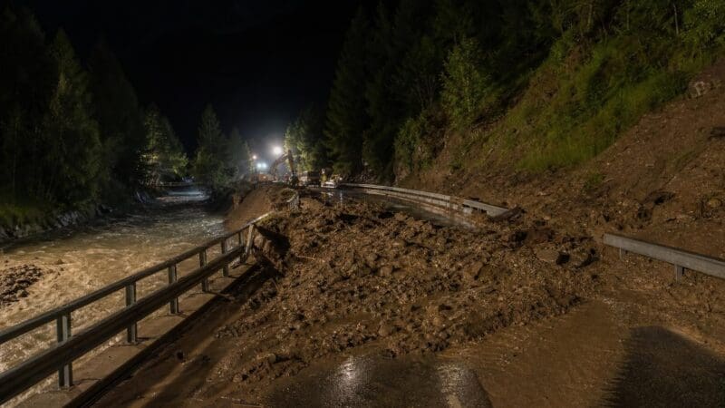 Eine von Schlamm und Geröll verschüttete Straße neben einem reißenden Fluss bei Nacht mit Einsatzkräften und Bagger.