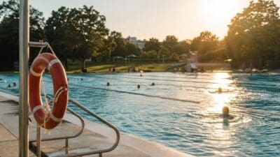 Roter Rettungsring am Geländer eines Freibads mit Schwimmern im Wasser bei Abendsonne