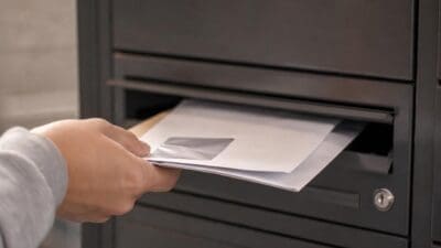 Eine Hand steckt mehrere weiße Briefe in den Einwurfschlitz eines dunklen Briefkastens.