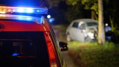 Rotes Einsatzfahrzeug mit blauem Blaulicht, im Hintergrund beschädigtes Auto an einem Baum