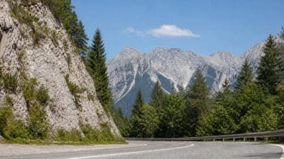 Geschwungene Bergstraße neben steiler Felswand mit Nadelbäumen und schneebedeckten Gipfeln