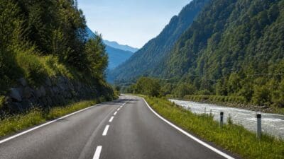 Zweispurige Asphaltstraße mit weißer Mittellinie führt durch grünes Bergtal neben Fluss und bewaldeten Hängen.