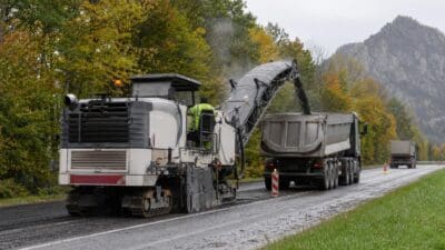 Große Straßenfräse bei der Belagssanierung auf einer mehrspurigen Straße mit Bäumen und Bergen im Hintergrund