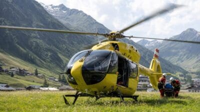 Gelber Rettungshubschrauber auf einer Wiese mit Rettungskräften und Trage vor Bergkulisse