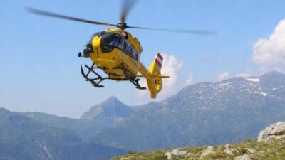 Gelber Rettungshubschrauber mit rot-weißer Heckmarkierung fliegt über Berglandschaft