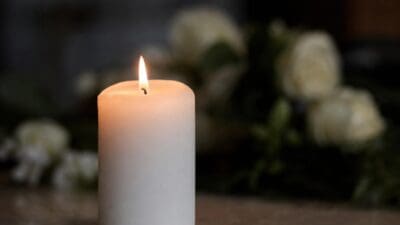 Eine brennende weiße Kerze steht auf einem Holztisch mit unscharfen weißen Blumen im Hintergrund