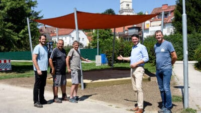 Vier Männer stehen neben einer Sandkiste mit rotem Sonnensegel auf dem Spielplatz in Wolfsberg.