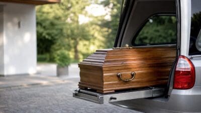 Dunkler Holzsarg wird aus dem Heck eines silbernen Bestattungsfahrzeugs auf Kopfsteinpflaster ausgefahren