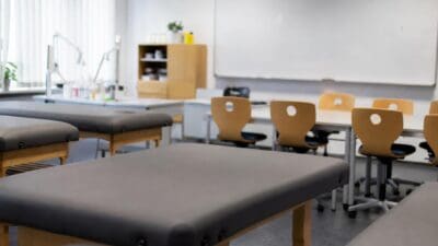 Leerer Klassenraum mit grauen Behandlungsliegen und Holzstühlen vor einem Whiteboard