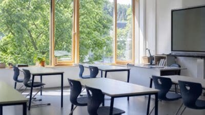 Heller Klassenraum mit Tischen und blauen Stühlen vor großen Fenstern mit Blick auf grüne Bäume