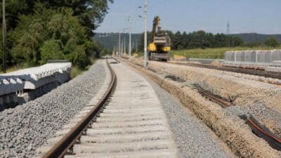 Blick auf eine Bahnstrecke mit neuen Betonschwellen und einem gelben Bagger im Hintergrund während Bauarbeiten