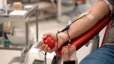 Nahaufnahme eines Arms bei der Blutspende mit Kanüle, Schlauch, Blutbeutel und rotem Herzball