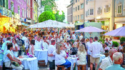 Menschen in weißer Kleidung sitzen an Tischen in einer belebten Innenstadt mit bunten Häusern und Sonnenschirmen.